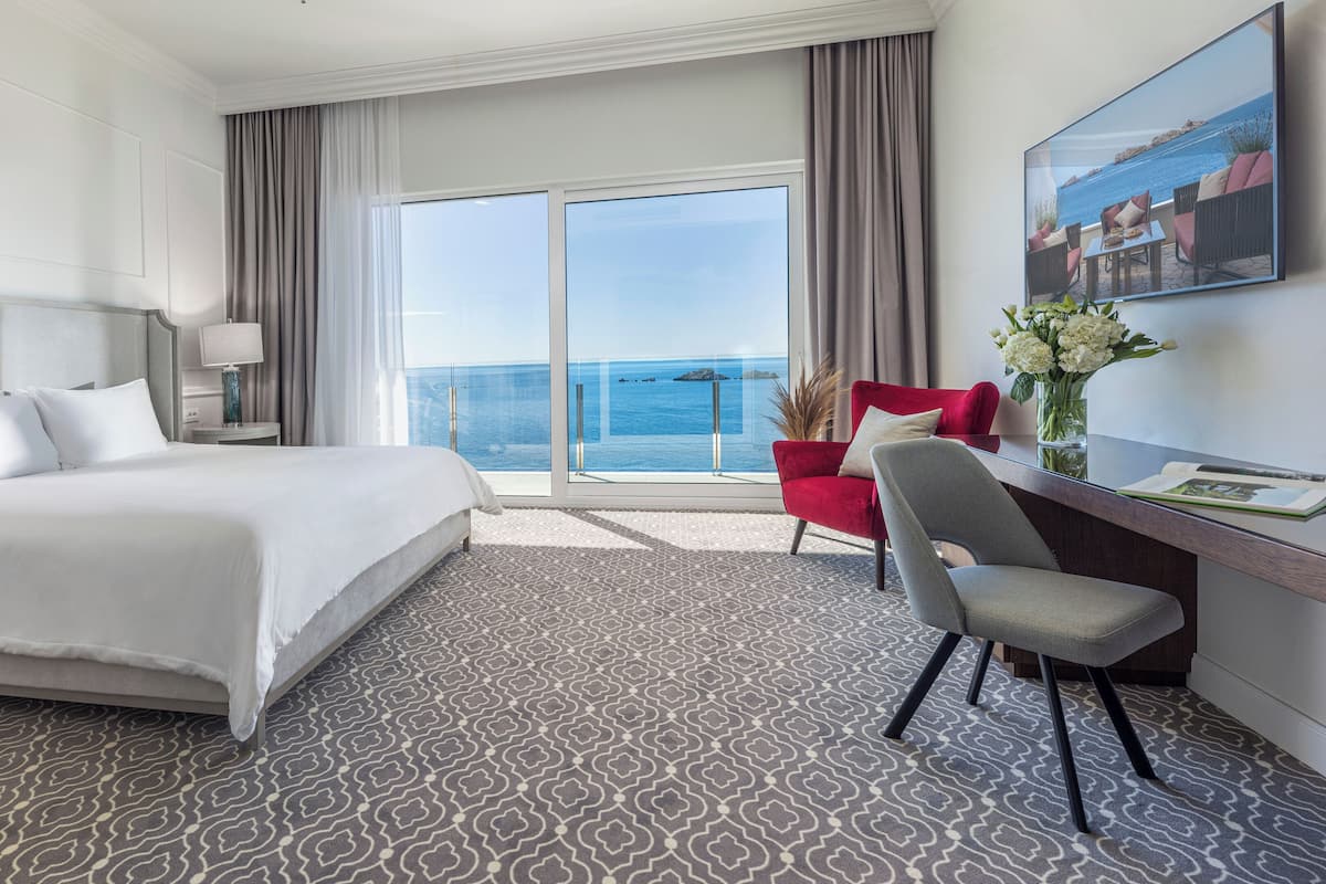 Quarto de hotel com uma cama grande, uma escrivaninha com cadeira, vista para o mar e um quadro na parede.