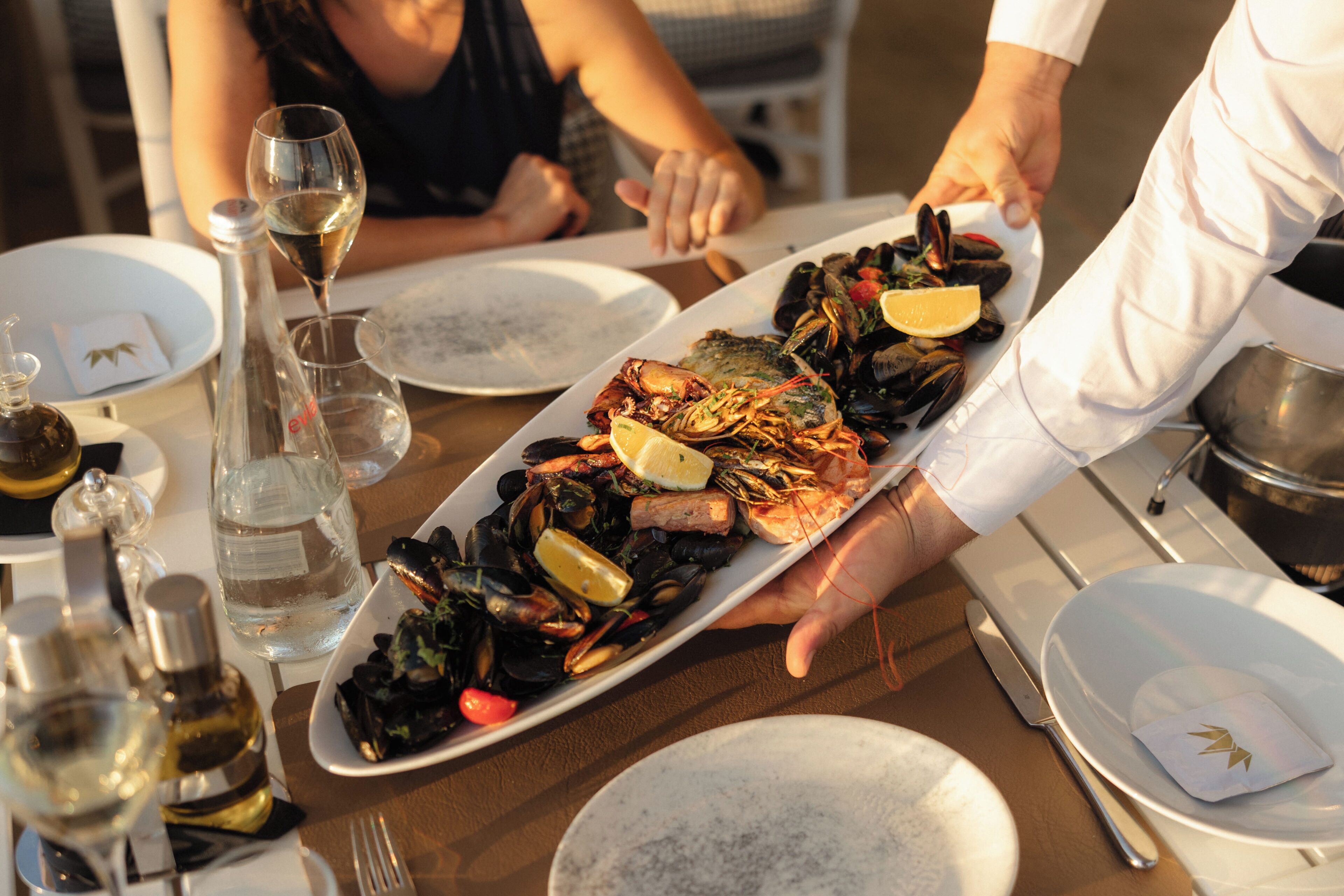 Uma seleção de mariscos sendo servida em uma mesa com copos de vinho e pratos.