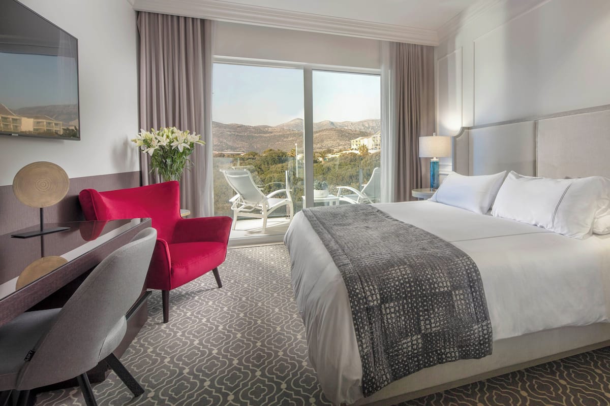Quarto de hotel com uma cama grande, uma secretária com cadeira, uma poltrona vermelha, vista para as montanhas pela janela e uma varanda com cadeiras.