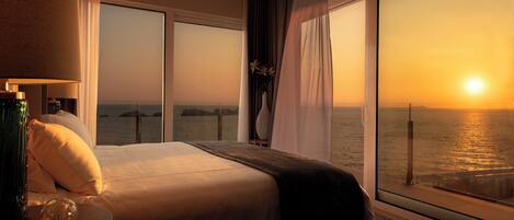Habitación de hotel con cama, mesita de noche, lámpara, jarra con flores y vista al océano al atardecer.