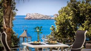 Un área de comedor al aire libre con una mesa dispuesta para dos, con vista a un mar azul y una costa rocosa.