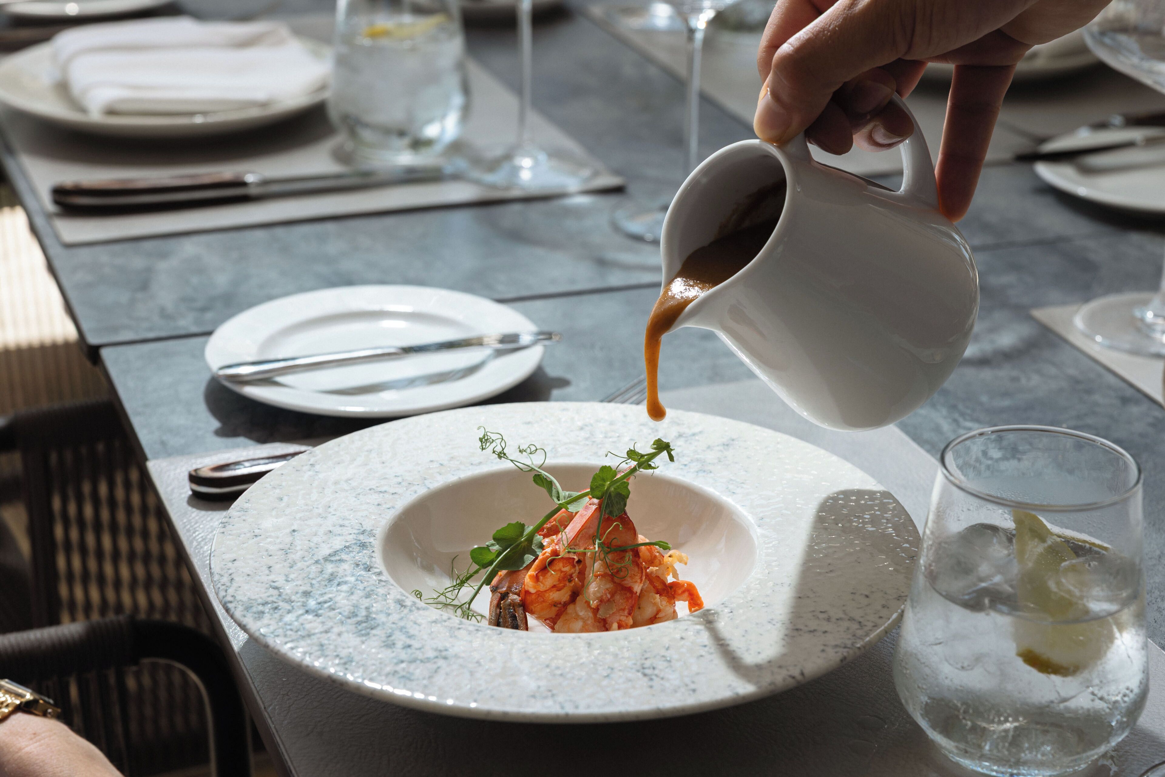 A dining table with a plate of food, a glass of water, and a hand pouring a sauce.