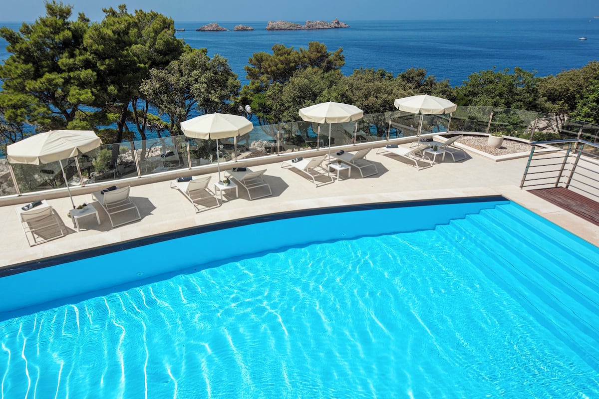 Área de piscina com espreguiçadeiras e guarda-sóis, com vista para o mar azul cristalino e a costa rochosa.