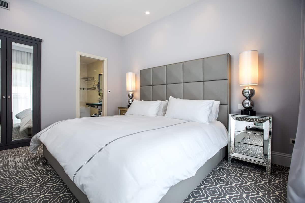 Quarto de hotel com uma cama grande, uma mesa de cabeceira com espelho e uma porta que dá para um banheiro.