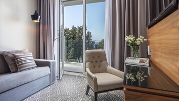 A modern hotel room with a grey sofa, a beige armchair, a wooden cabinet, and a view of trees and a body of water through the glass door.