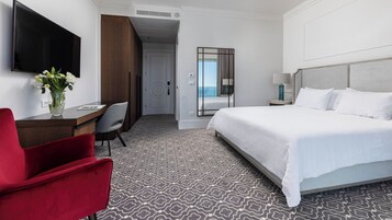 Quarto de hotel com cama, escrivaninha com cadeira, televisão e vista para o oceano.