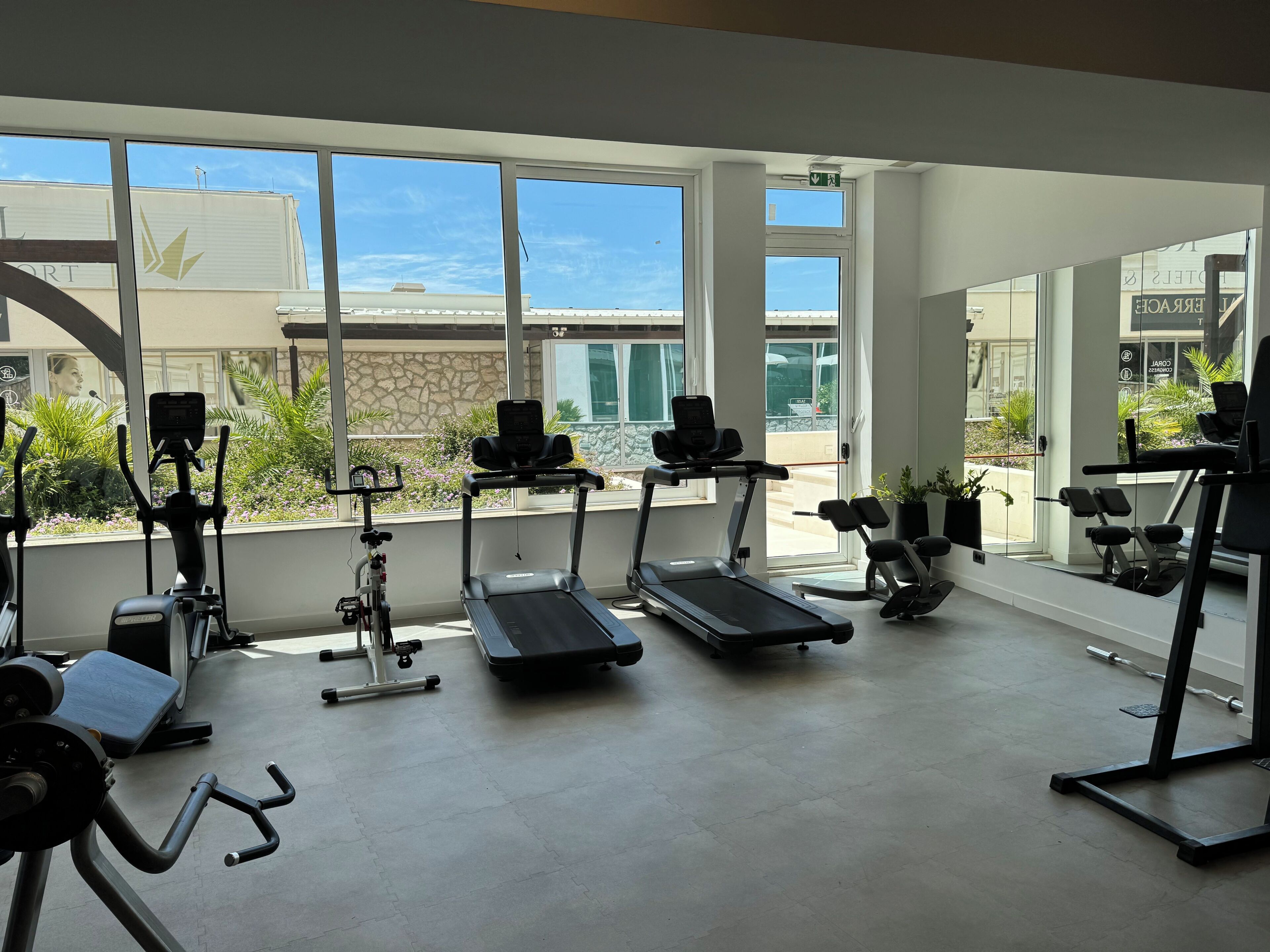 Uma academia com esteiras, elípticos e equipamentos de musculação. Grandes janelas oferecem vista para o exterior.