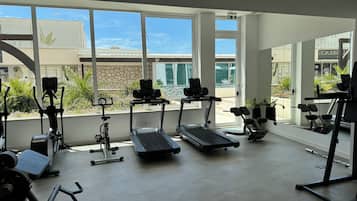 Uma academia com esteiras, elípticos e equipamentos de musculação. Grandes janelas oferecem vista para o exterior.