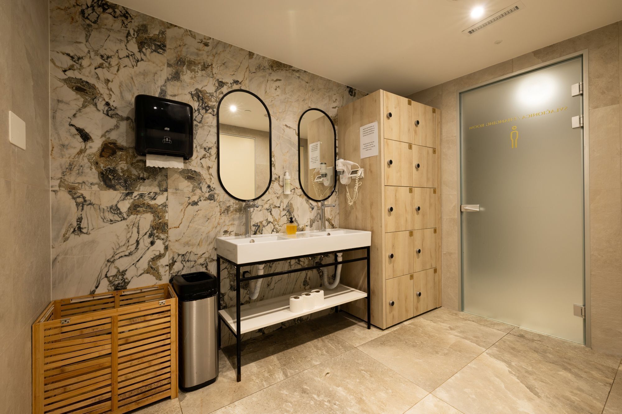 A modern bathroom with a marble backsplash, a white sink, a mirror, a wooden cabinet, and a trash bin.