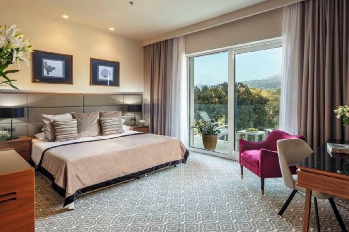 Habitación de hotel con cama, escritorio, silla, ventana con cortinas y balcón con vistas.