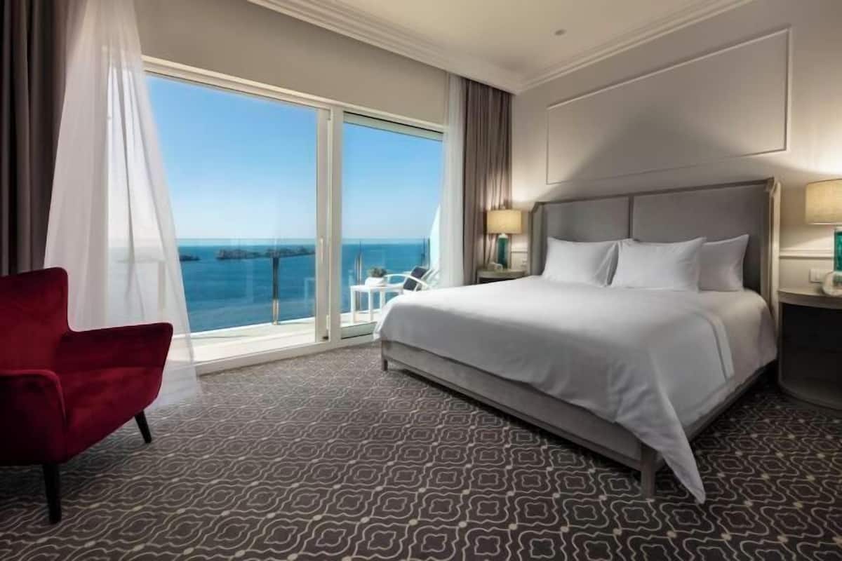 A hotel room with a large bed, a red chair, a balcony with a view of the sea, and a patterned carpet.