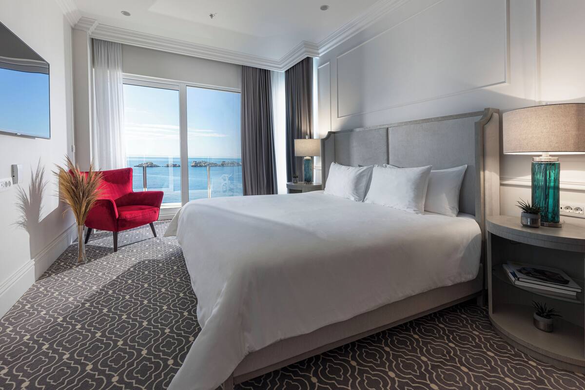 A hotel room with a large bed, a red armchair, a nightstand with a lamp, and a view of the sea.