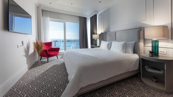 Quarto de hotel com uma cama grande, uma poltrona vermelha, uma mesa de cabeceira com luminária e vista para o mar.