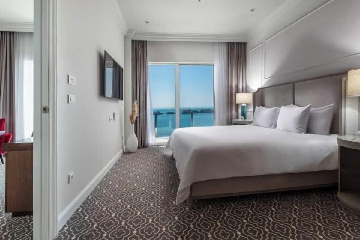 A hotel room with a large bed, a television, a nightstand with a lamp, and a view of the sea through the window.