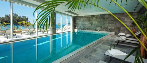 A modern indoor swimming pool with clear blue water, surrounded by lounge chairs and a view of the ocean.