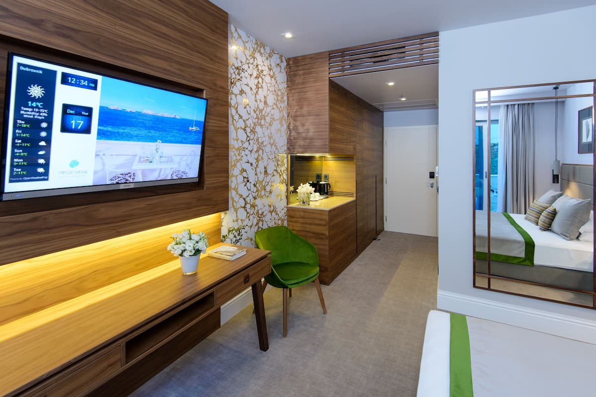 A modern hotel room with a wooden desk, a green chair, a bed with a green bedspread, a large mirror, and a TV displaying the weather.