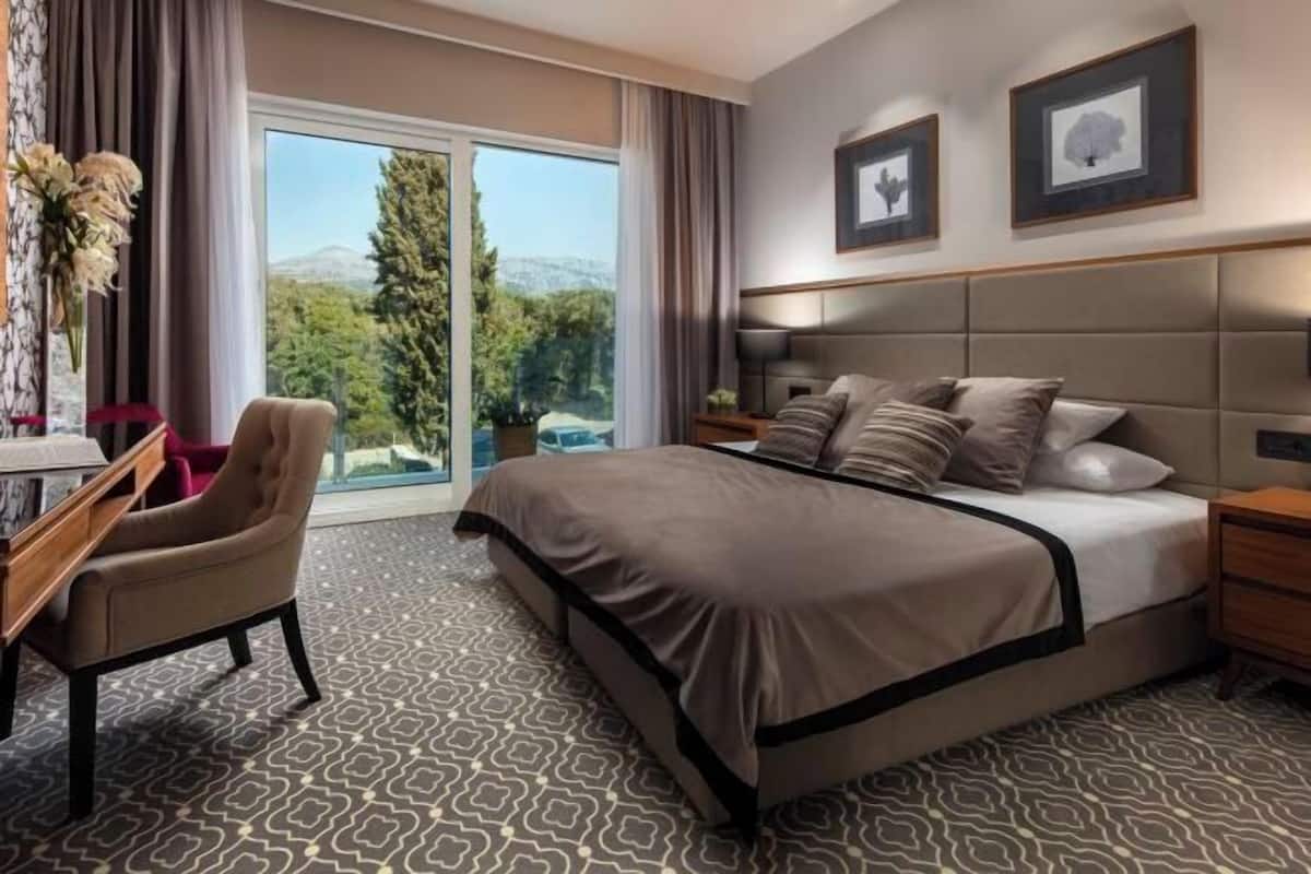 A hotel room with a large bed, a desk, a chair, a nightstand, and a view of the outdoors.