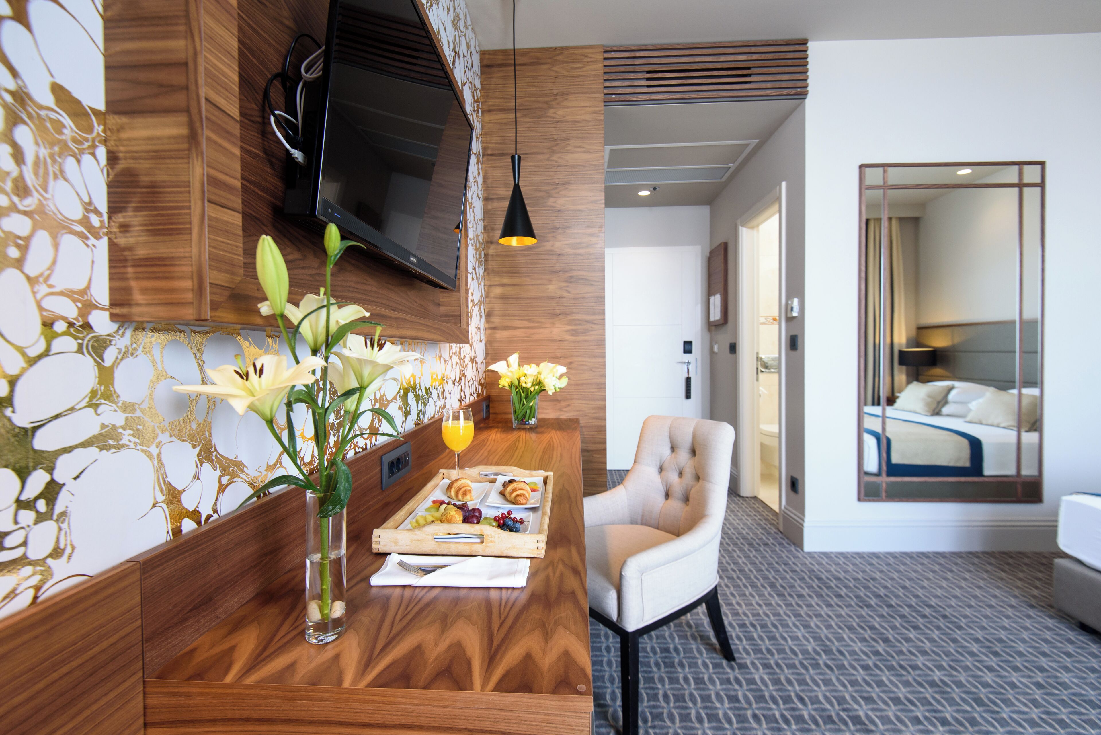 A hotel room with a wooden reception desk, a comfortable armchair, a bed with a view, and a TV mounted on the wall.