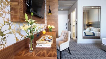 A hotel room with a wooden reception desk, a comfortable armchair, a bed with a view, and a TV mounted on the wall.