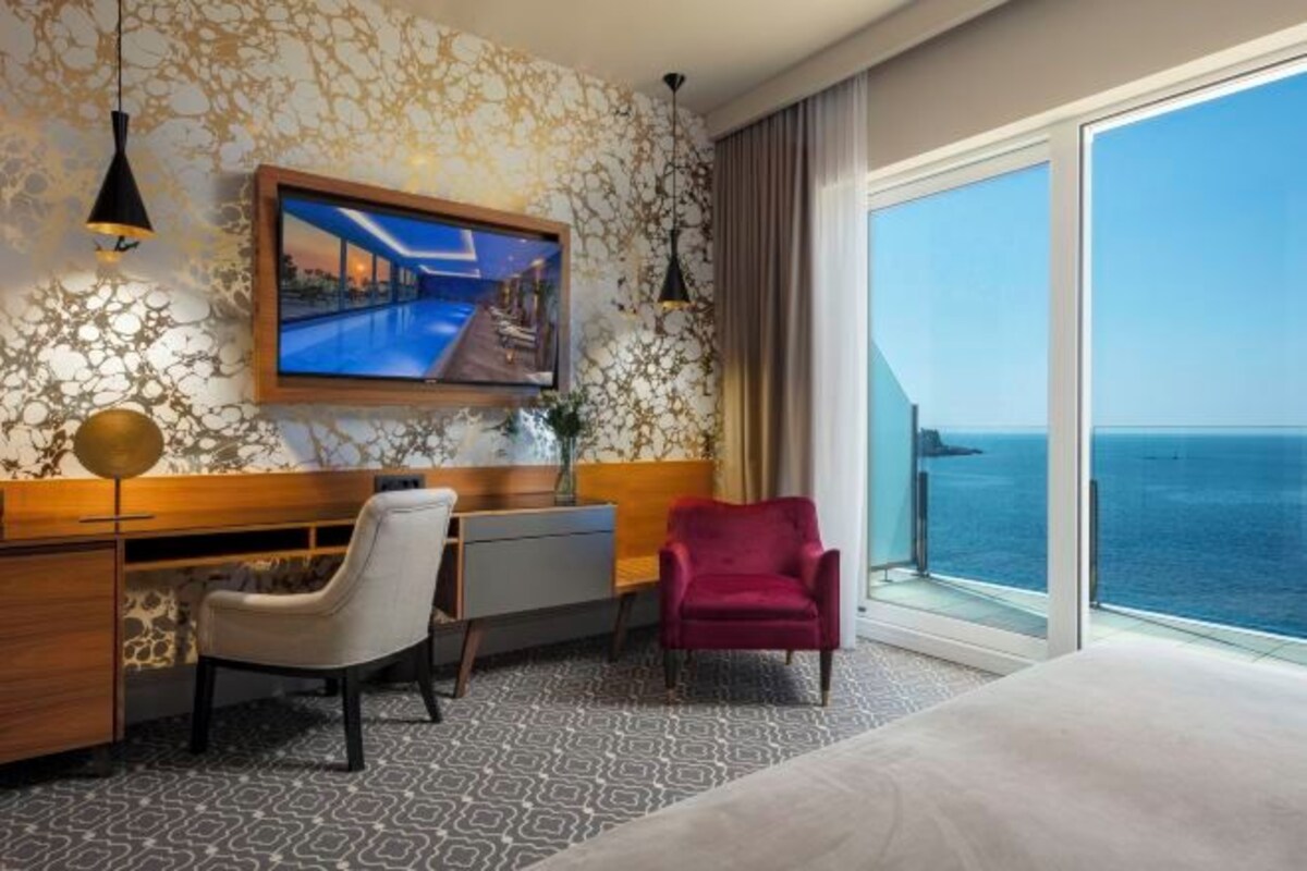 Habitación de hotel con una televisión grande, un escritorio, una silla, un sillón rojo y vista al mar.