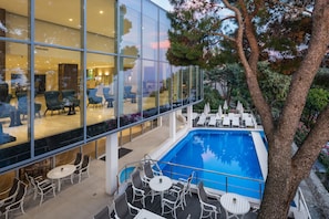 Um hotel moderno com fachada de vidro, área de piscina com espreguiçadeiras e espaço de refeições com mesas e cadeiras.