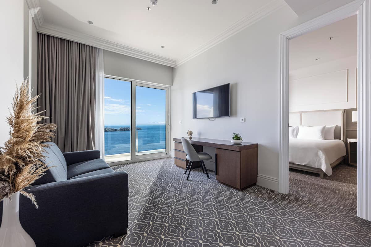 A hotel room with a large window, a desk, a bed, and a view of the sea.