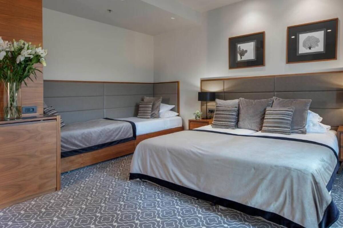 A hotel room with two single beds, a large bed, a wooden nightstand, a bedside table, and a vase with flowers.