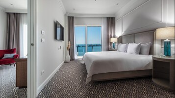 A hotel room with a large bed, a red armchair, a nightstand with a lamp, and a view of the sea through floor-to-ceiling windows.