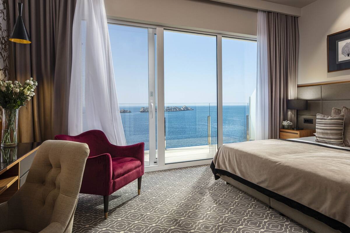 A hotel room with a large bed, a red chair, a desk with a lamp, and a view of the sea through a glass door.