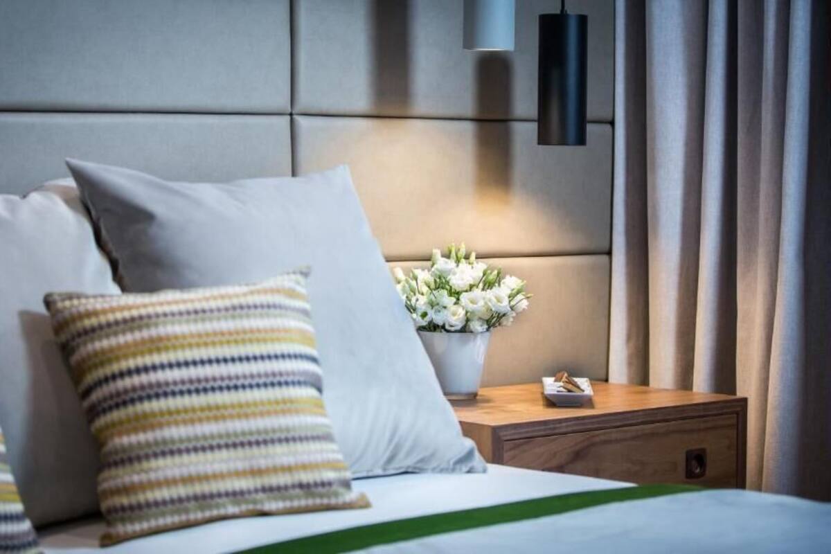 A hotel room with a bed, pillows, a nightstand, a vase with flowers, and a lamp.