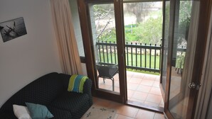 Een woonkamer met een donkere gestreepte bank, een bijzettafeltje en een schuifdeur naar een balkon met een stoel en uitzicht op groen.