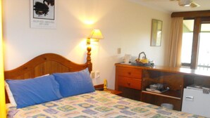 Een hotelkamer met een houten bed, een nachtkastje, een bureau met lamp en een spiegel, plus een raam met gordijnen.