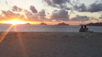 A beach with a sunset, two people sitting on the sand, and distant islands.