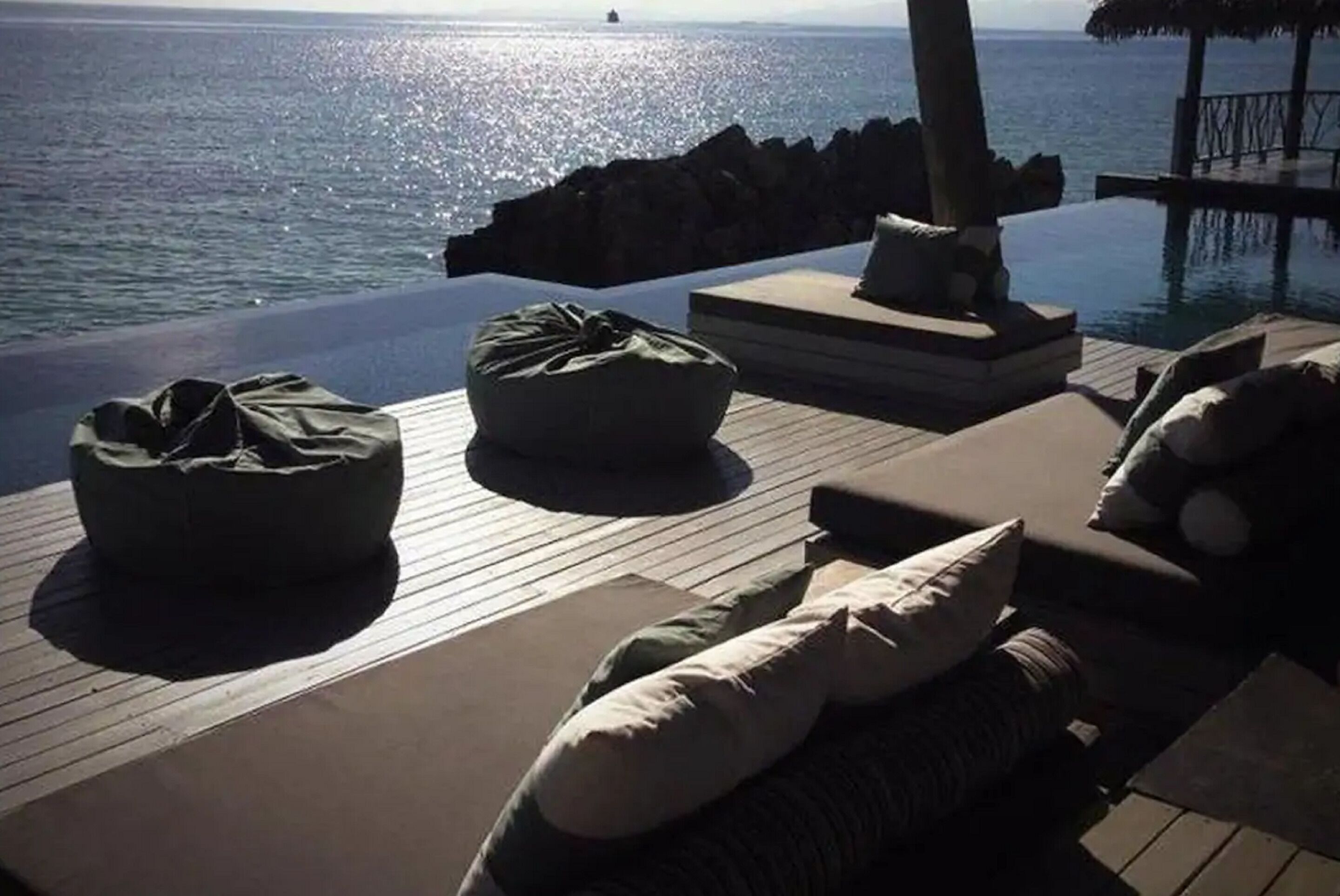 An infinity pool with a view of the ocean, wooden deck seating, and a thatched-roof structure.