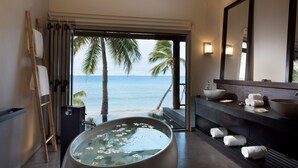 A modern bathroom with a large bathtub, a sink, and a mirror. There are towels on the floor and a view of the ocean through an open door. - Tadrai Island Resort-Fiji - All Inclusive (Mana Island)