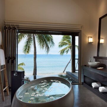 A modern bathroom with a large bathtub, a sink, and a mirror. There are towels on the floor and a view of the ocean through an open door.