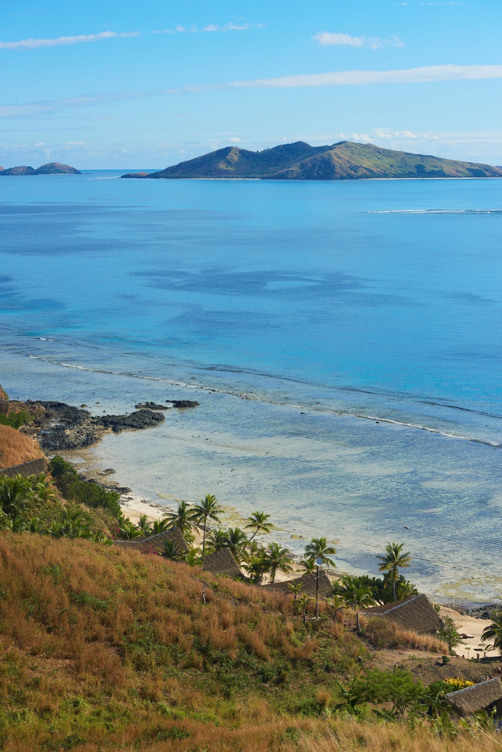 A coastal landscape with a beach, palm trees, and distant hills.