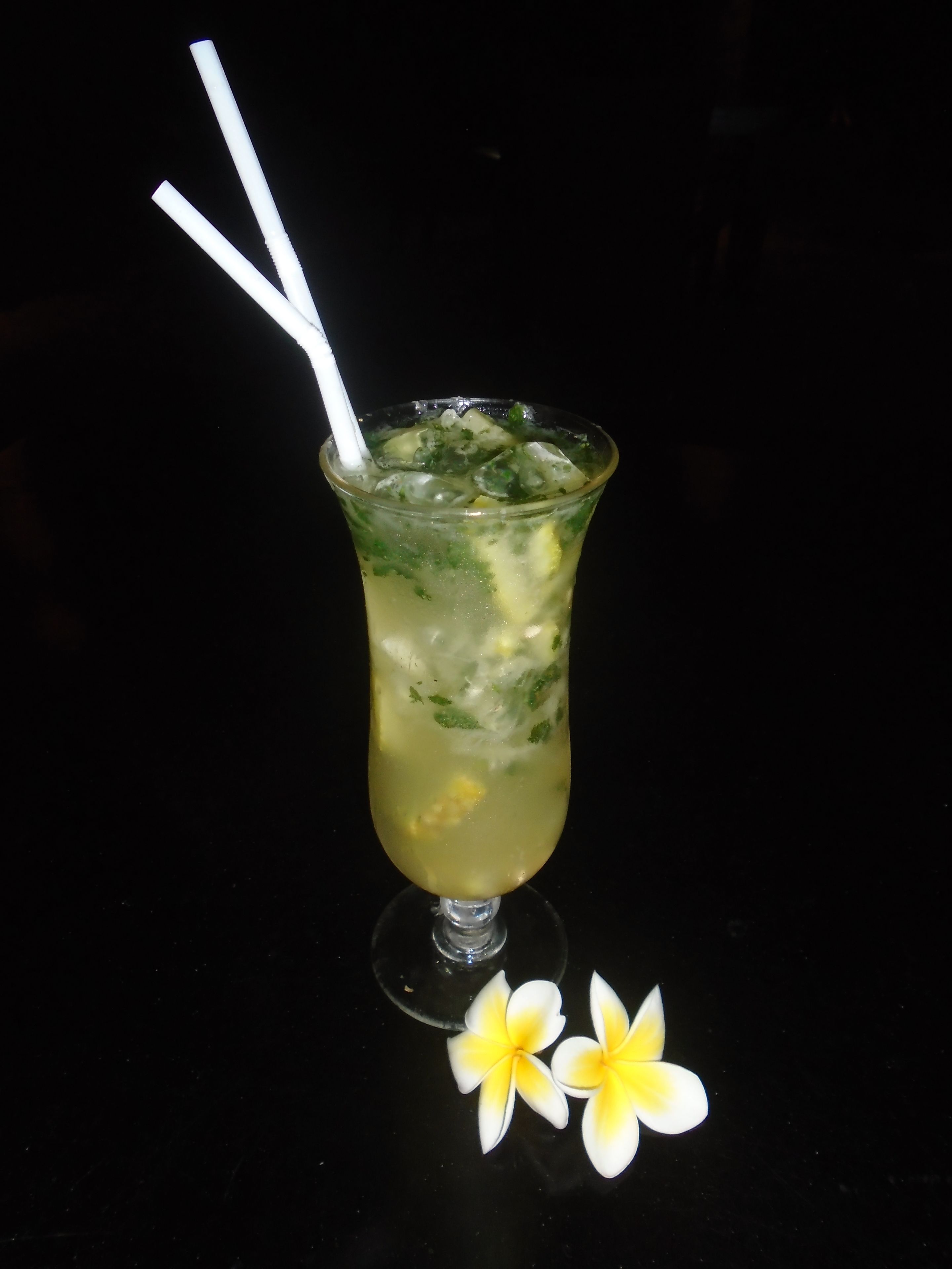 A cocktail glass with a straw, ice, and garnishes, accompanied by two plumeria flowers on a black surface.
