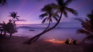 A beach scene with palm trees, a fire, and two people sitting on the sand. - Tadrai Island Resort-Fiji - All Inclusive (Mana Island)