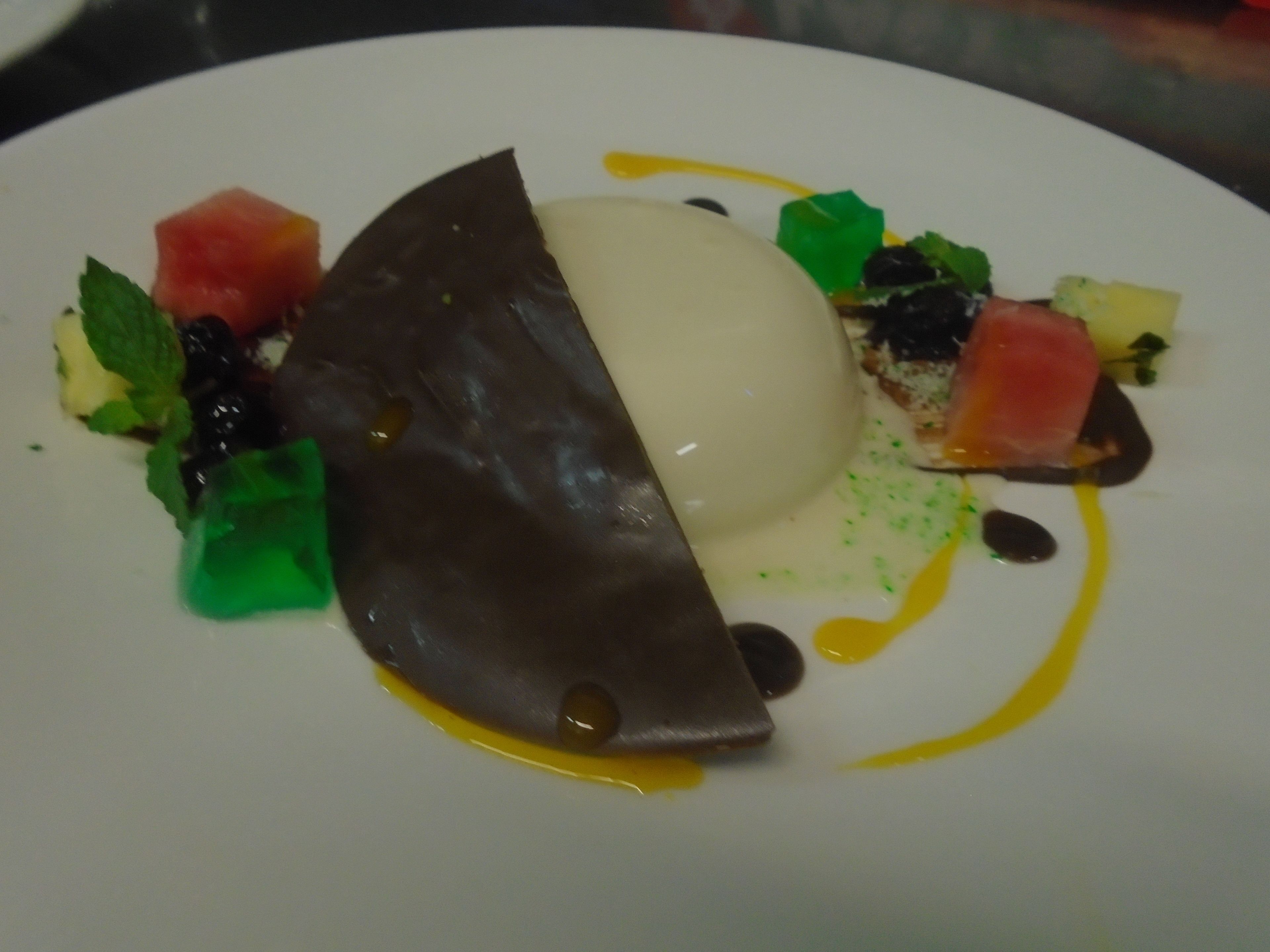 Un dessert con una cupola di cioccolato, una pallina di gelato e guarnizioni colorate su un piatto bianco.