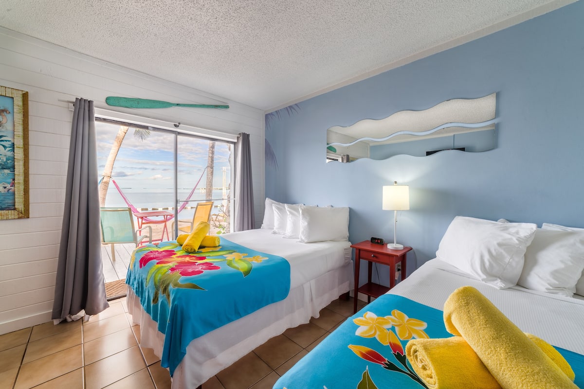 A hotel room with two beds, a window with curtains, a chair outside, and a view of the beach.