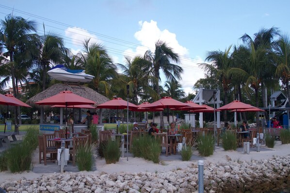 Outdoor seating with red umbrellas, palm trees, and a jet ski in the background.