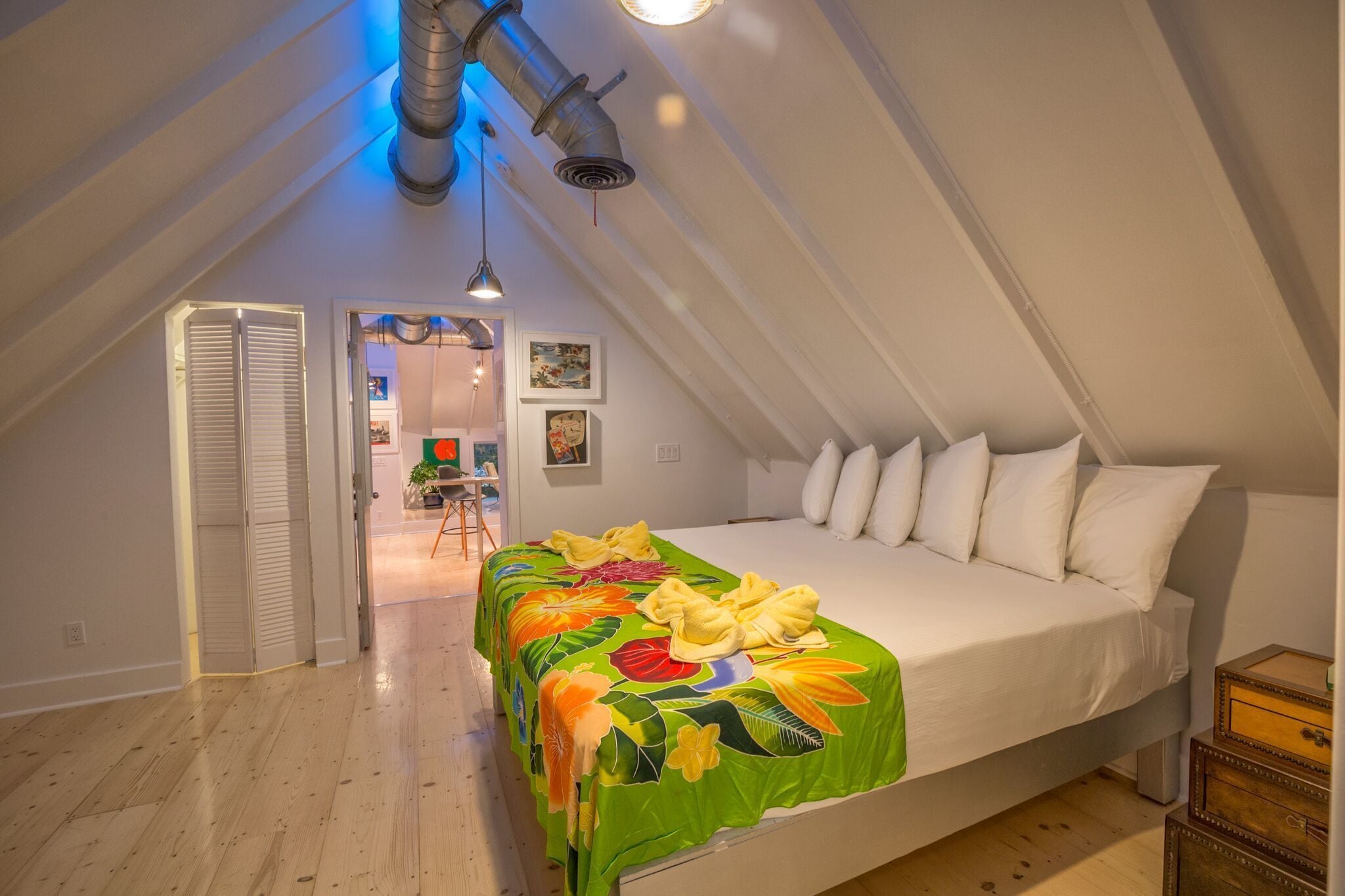 Een slaapkamer met een bed, witte kussens en een beddegoed met groene bloemen. Er ligt een houten vloer en het plafond heeft leidingwerk.