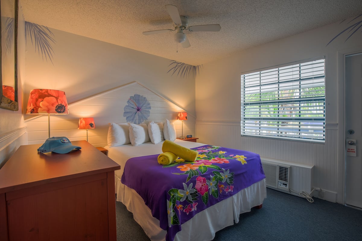 A hotel room with a bed, two bedside lamps, a ceiling fan, a window with blinds, and a wooden nightstand with a cap.