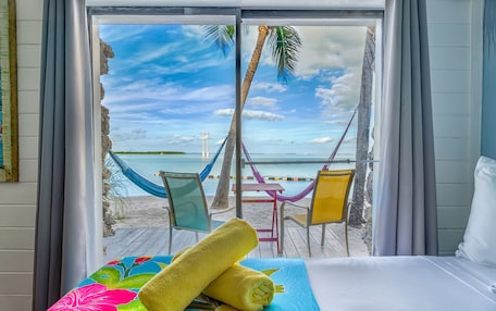 A hotel room with a bed, a view of the beach, a hammock, and beach chairs.