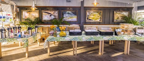 A buffet setup with various food items and drinks, surrounded by tropical-themed decorations and fish paintings on the wall.