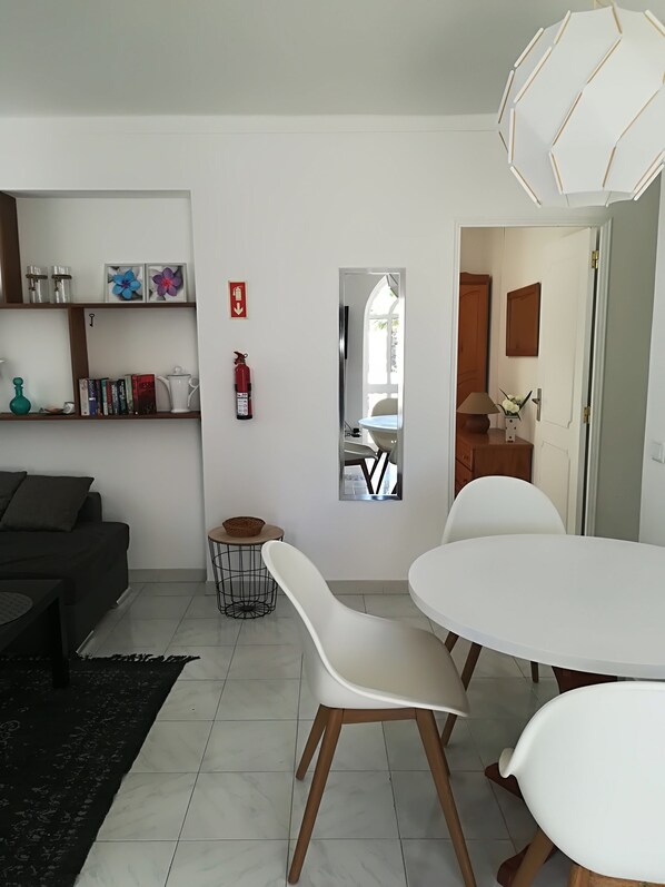 Un salon moderne avec un canapé blanc, une table à manger entourée de chaises blanches et une cuisine visible à travers une fenêtre.