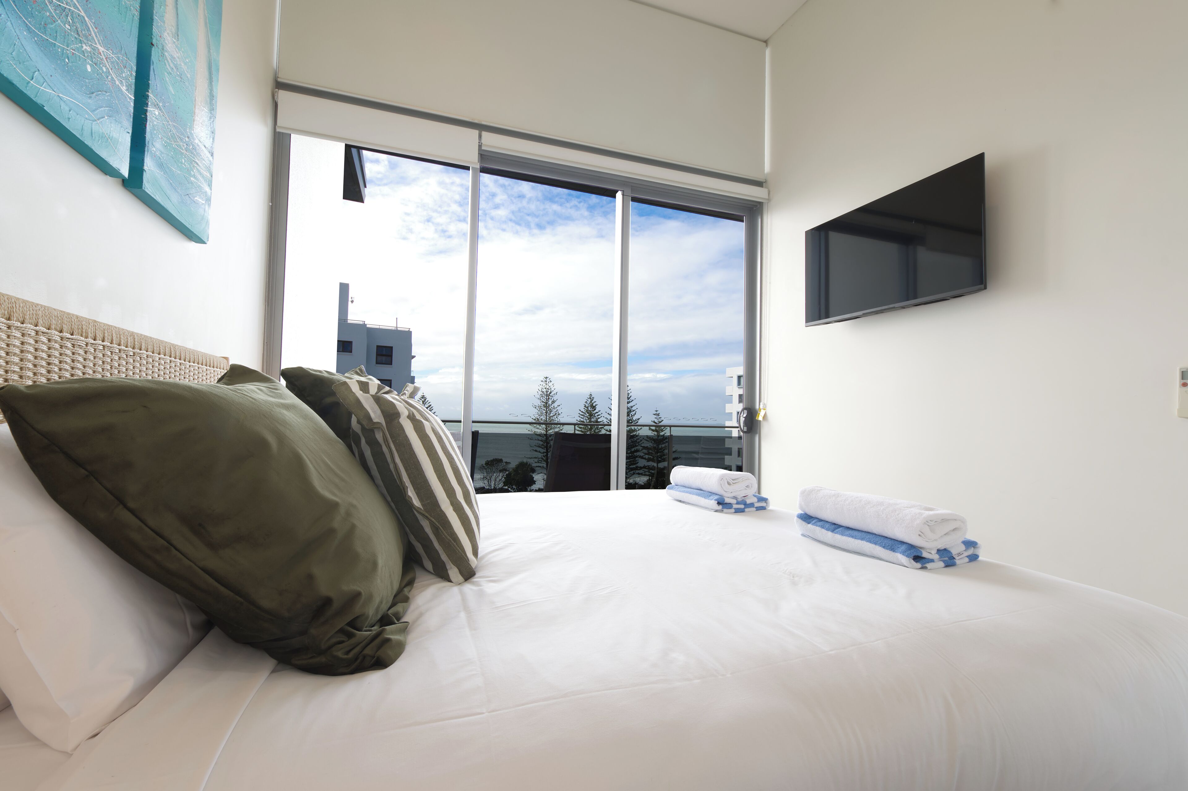A hotel room with a large bed, a flat-screen TV mounted on the wall, and a view of the city through large windows.