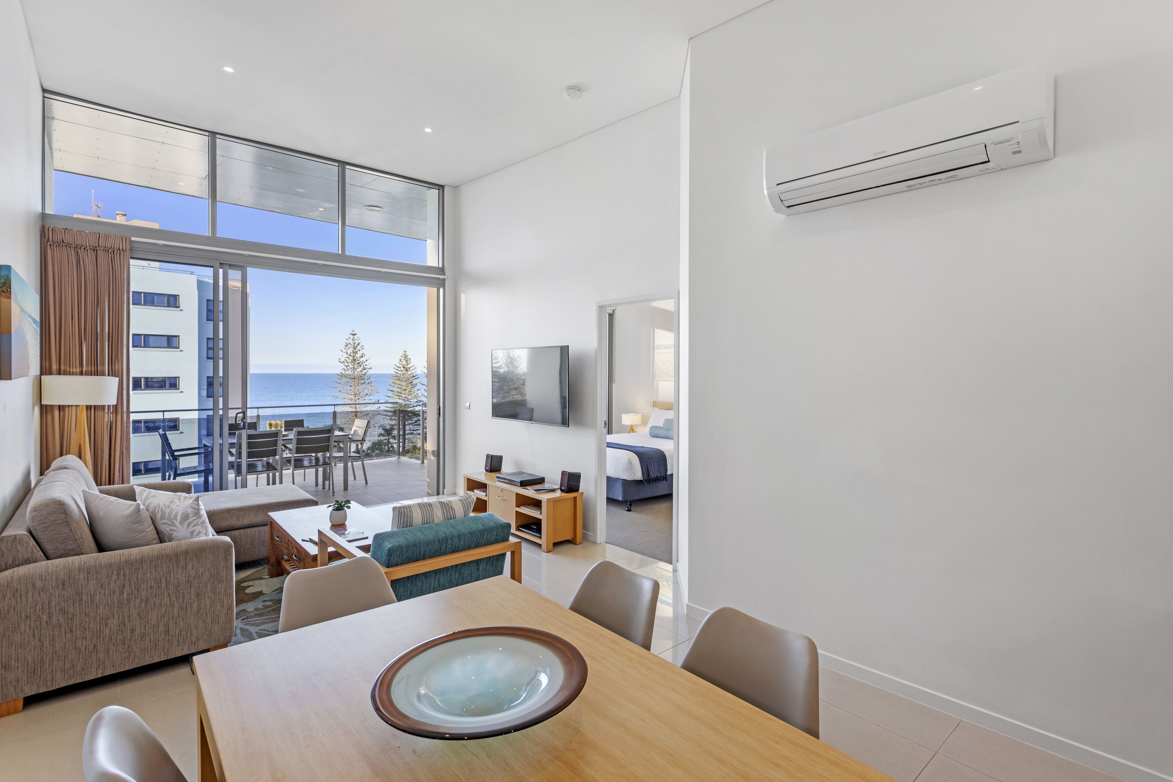 A modern hotel room with a dining area, a sofa, a TV, and a balcony with a view of the sea.