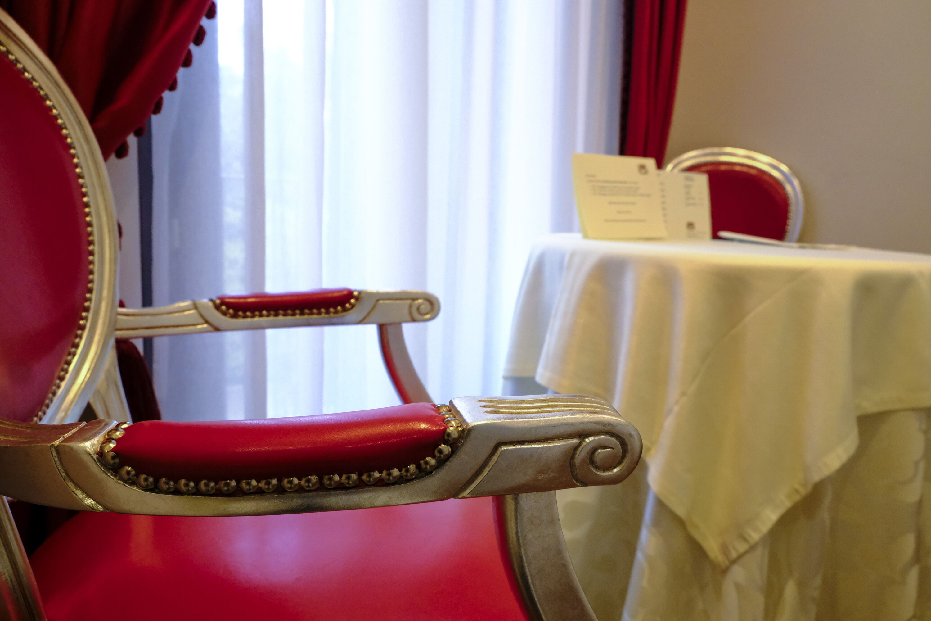 A red upholstered chair with a gold frame, a white table with a white tablecloth, and a window with white curtains in the background.